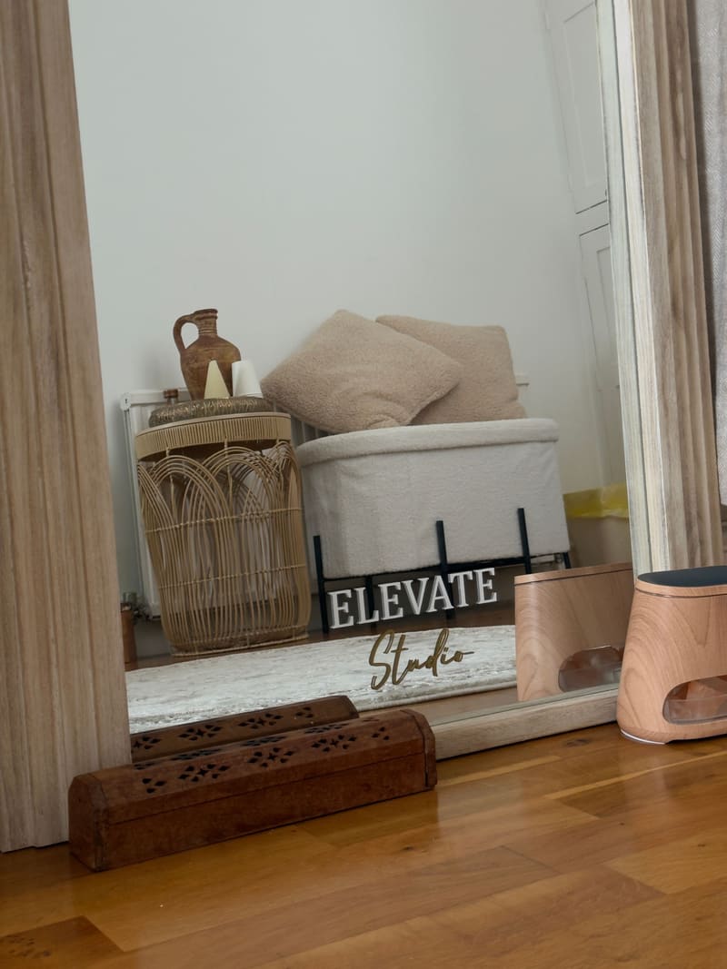 An ‘Elevate Studio’ sign reflected in a tall floor mirror beside a rattan side table and bouclé cushions