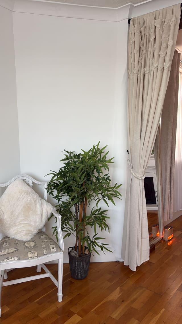 A white chair with a sheepskin cushion beside a leafy plant and linen curtains