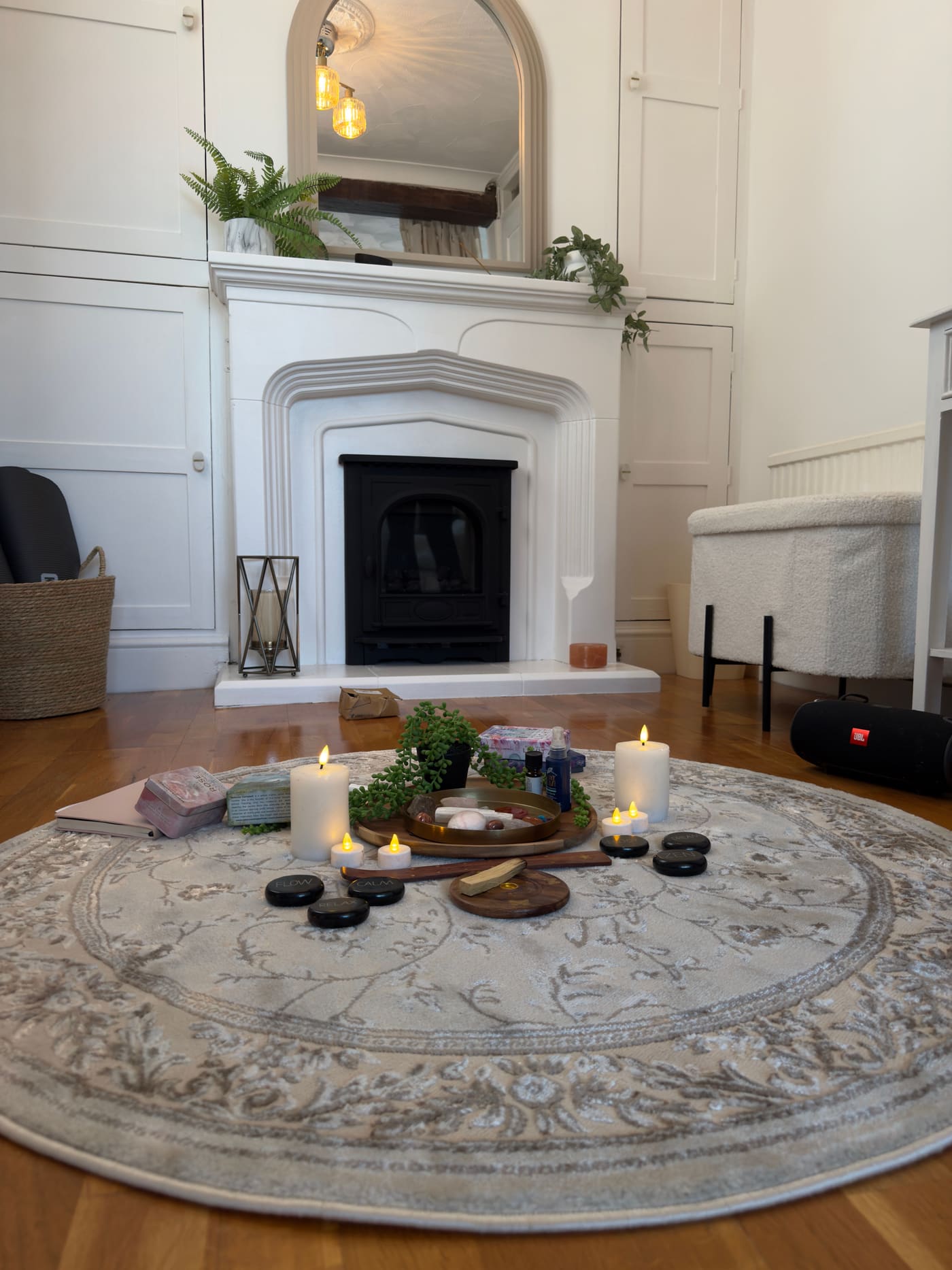 Candles, crystals and smooth stones arranged on a round rug in front of the studio’s white fireplace and arched mirror