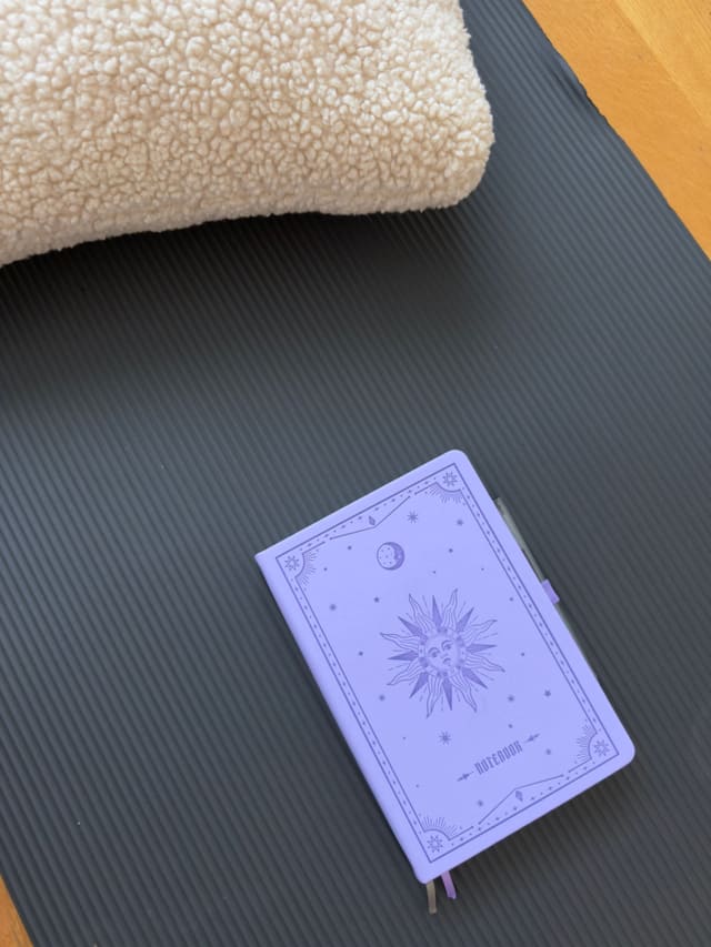 A notebook with a sun and moon design resting on a yoga mat beside a soft bouclé cushion